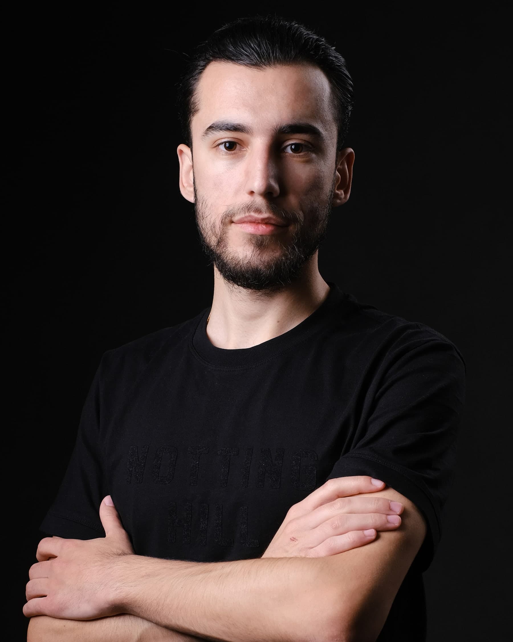 Portrait of Orgito Baraj with arms crossed, wearing a black t-shirt against a dark background