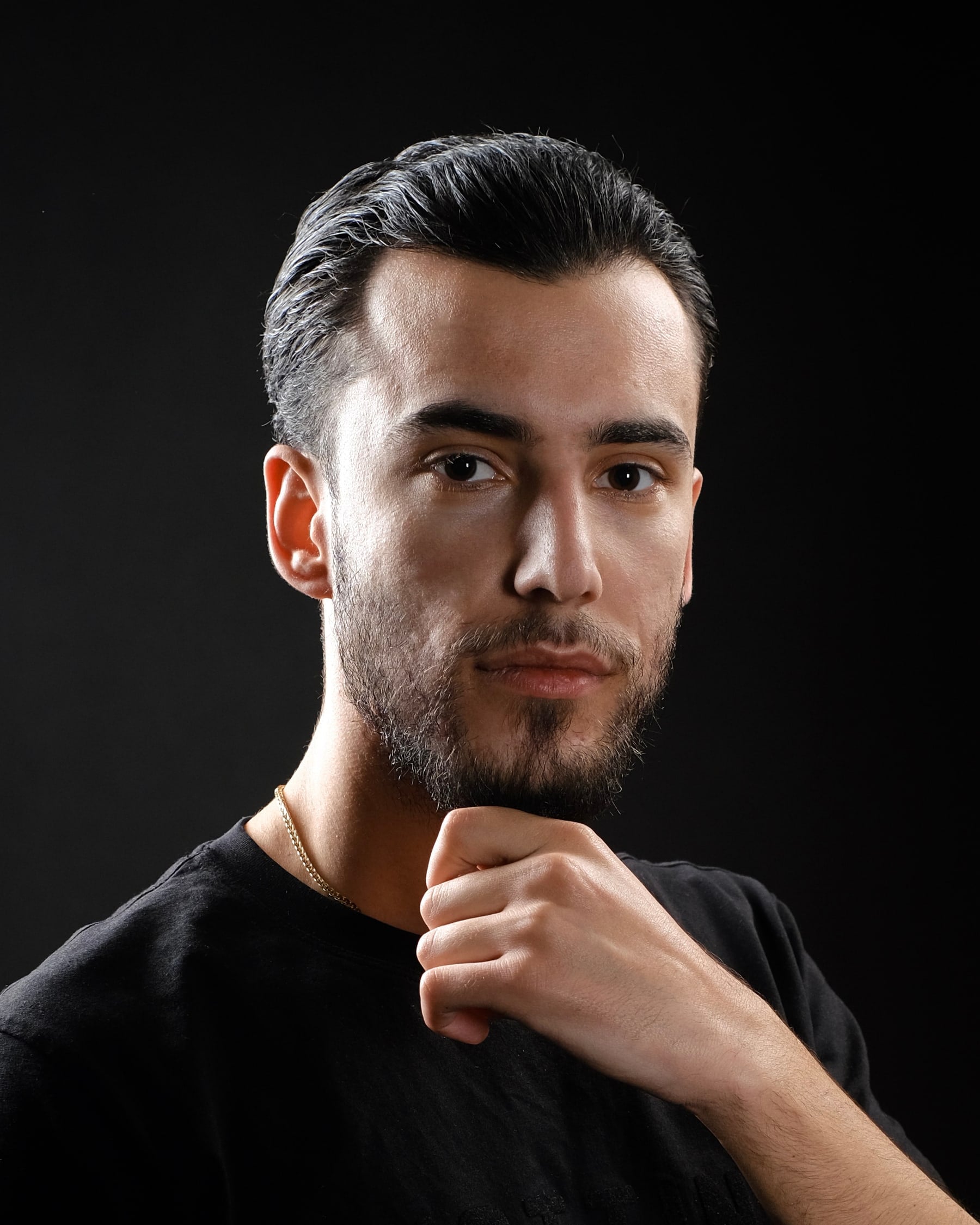 Portrait of Orgito Baraj resting his chin on his hand, wearing a black t-shirt against a dark background